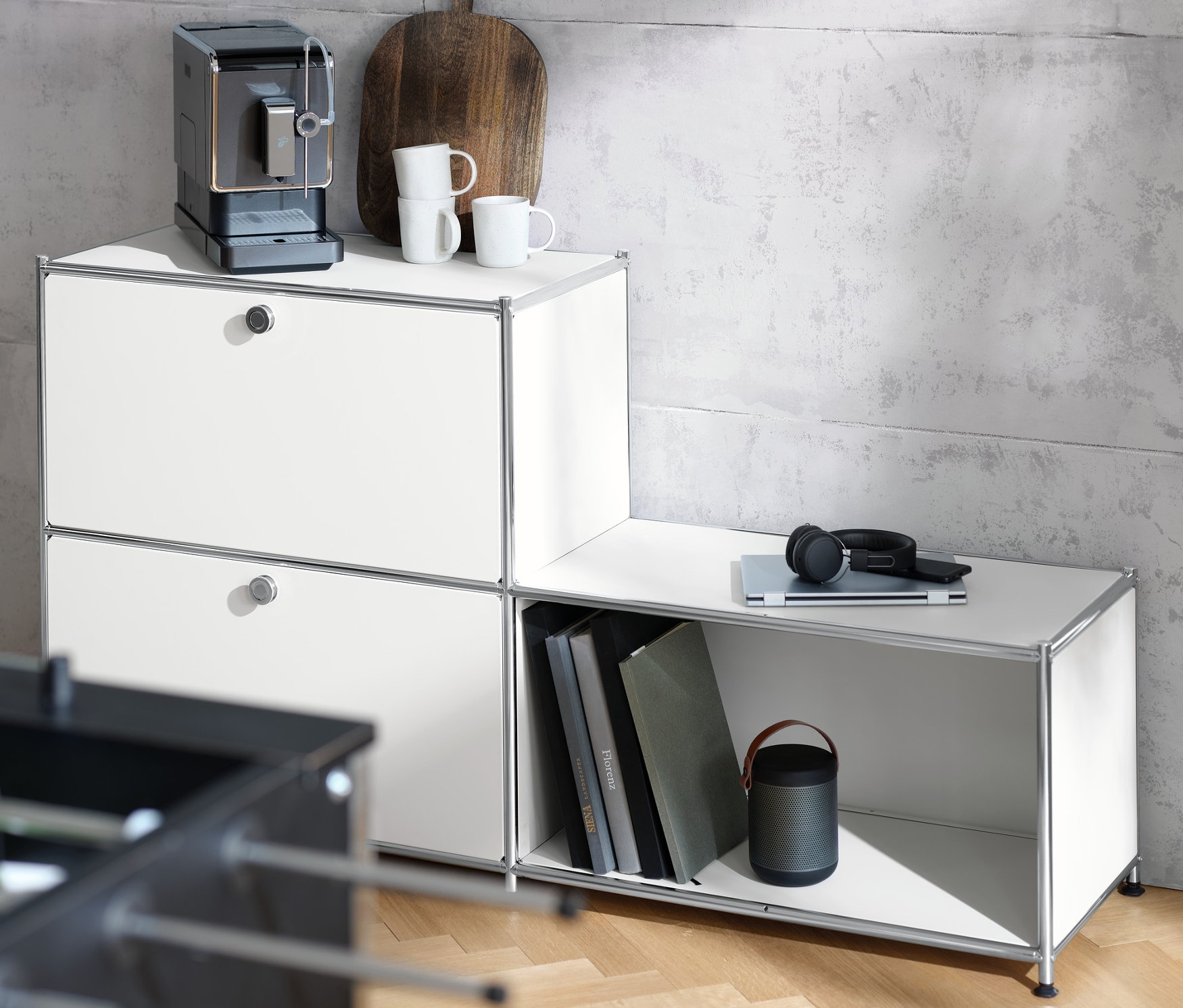 Ekspres do kawy stoi na białym metalowym regale »CN3« z 3 półkami. Na regale leżą również filiżanka, deska i słuchawki.