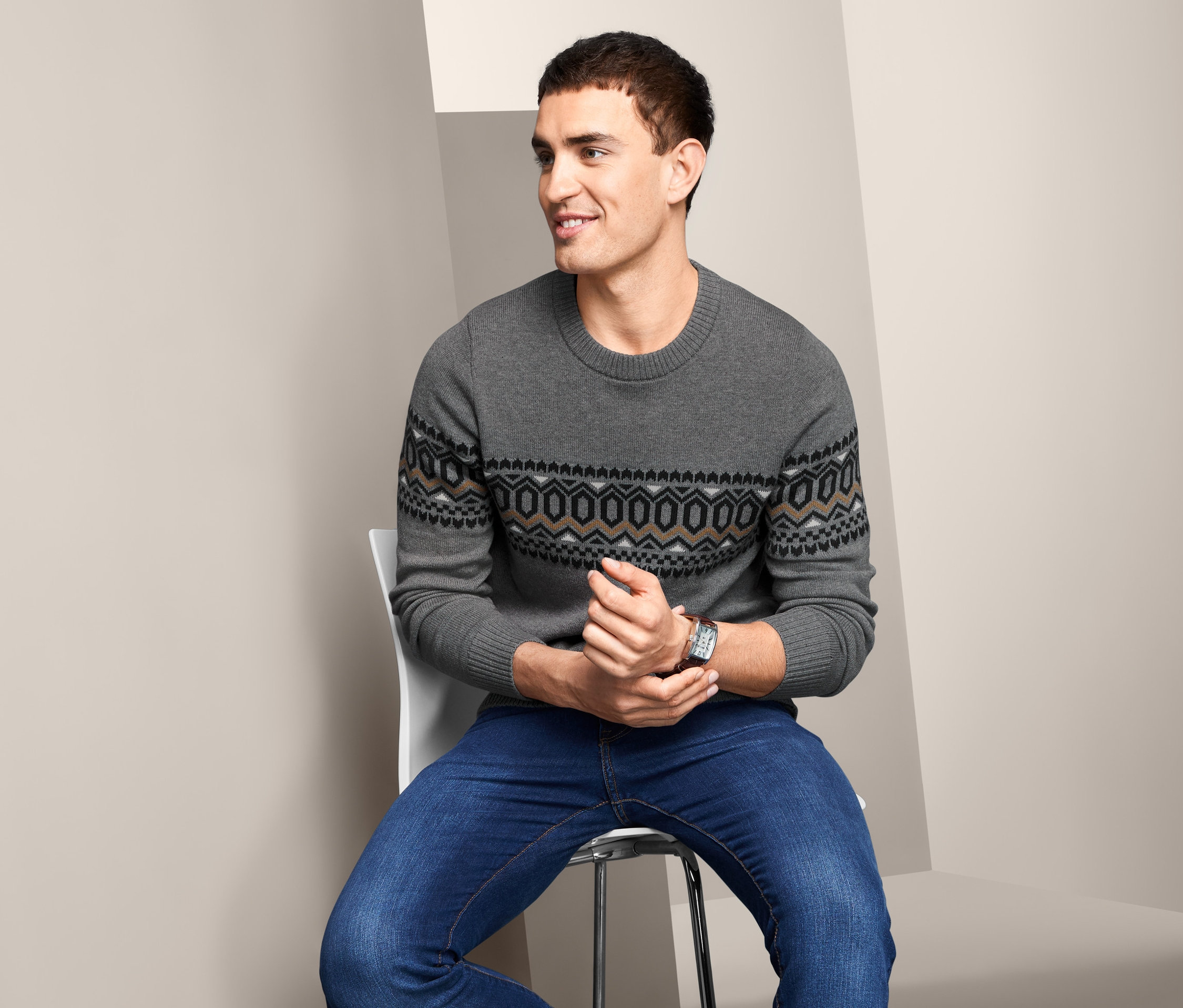 Mężczyzna siedzi na krześle i ma na sobie szary sweter z okrągłym wycięciem pod szyją i dżinsy.