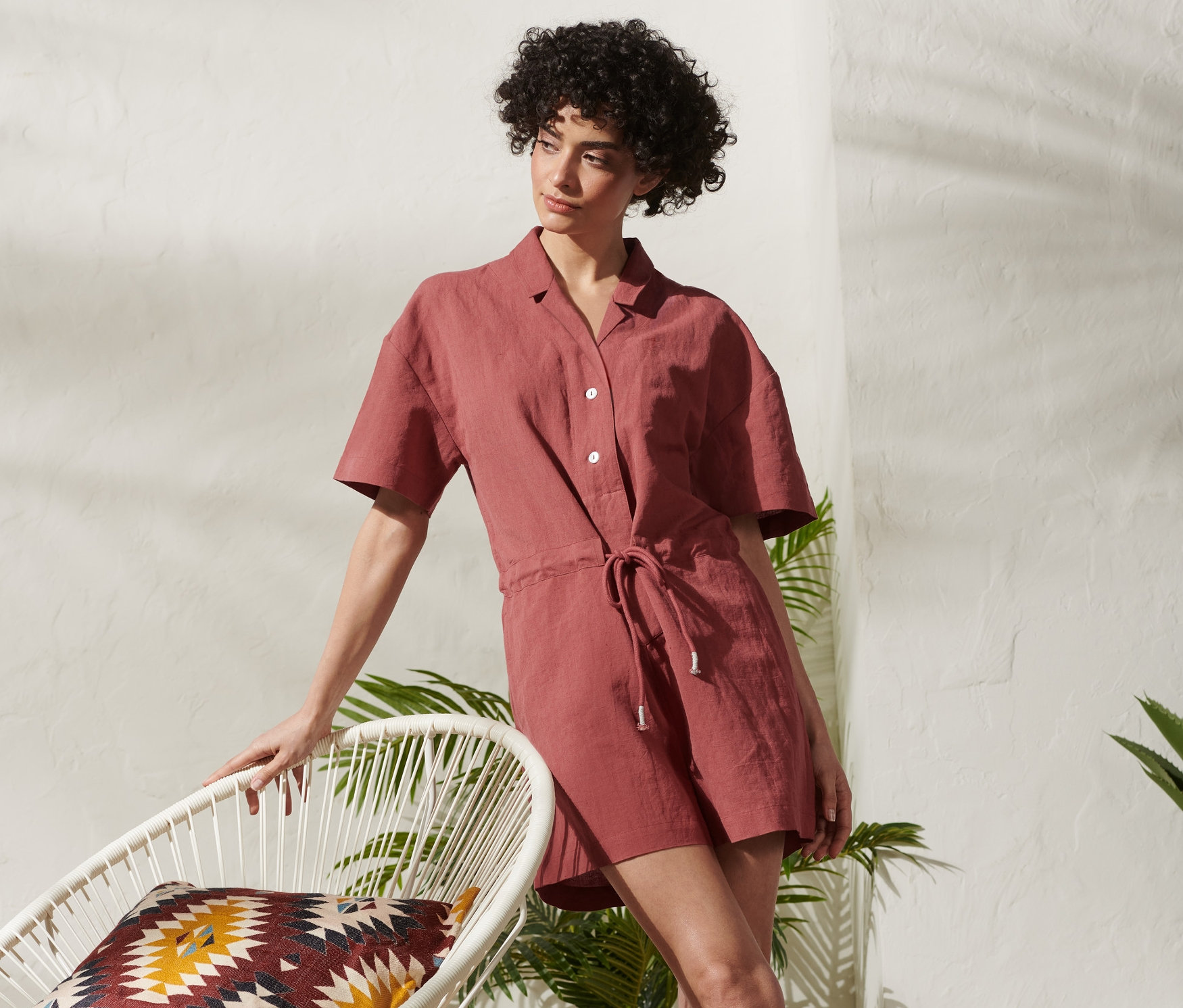 Kobieta opiera się o białe krzesło w krótkim kombinezonie z lnem.