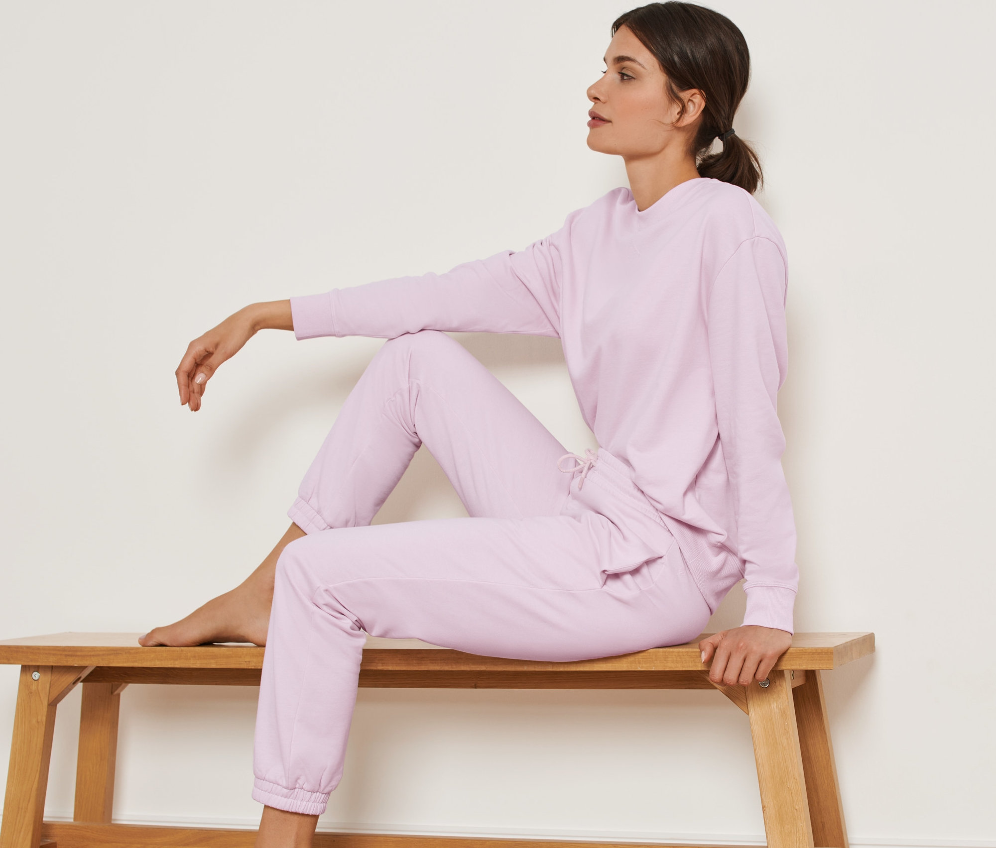 Kobieta siedzi na drewnianej ławce i ma na sobie spodnie dresowe w kolorze jasnego różu.