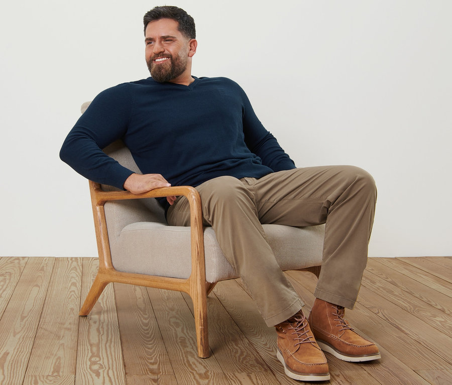 Mężczyzna siedzi w fotelu ubrany w piaskowe spodnie chino i granatowy sweter z dekoltem w serek.