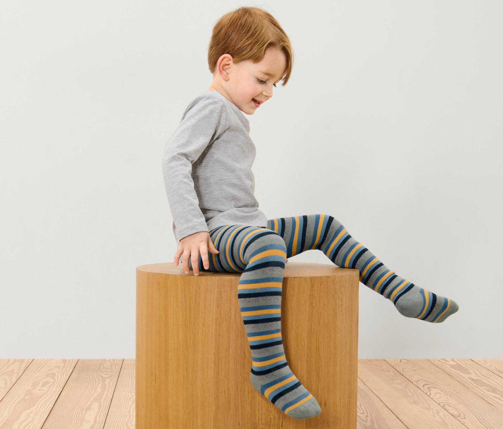 Chłopiec siedzi na drewnianym stołku i ma na sobie dziecięce rajstopy termoaktywne, 2 pary, niebieskie i wielokolorowe.