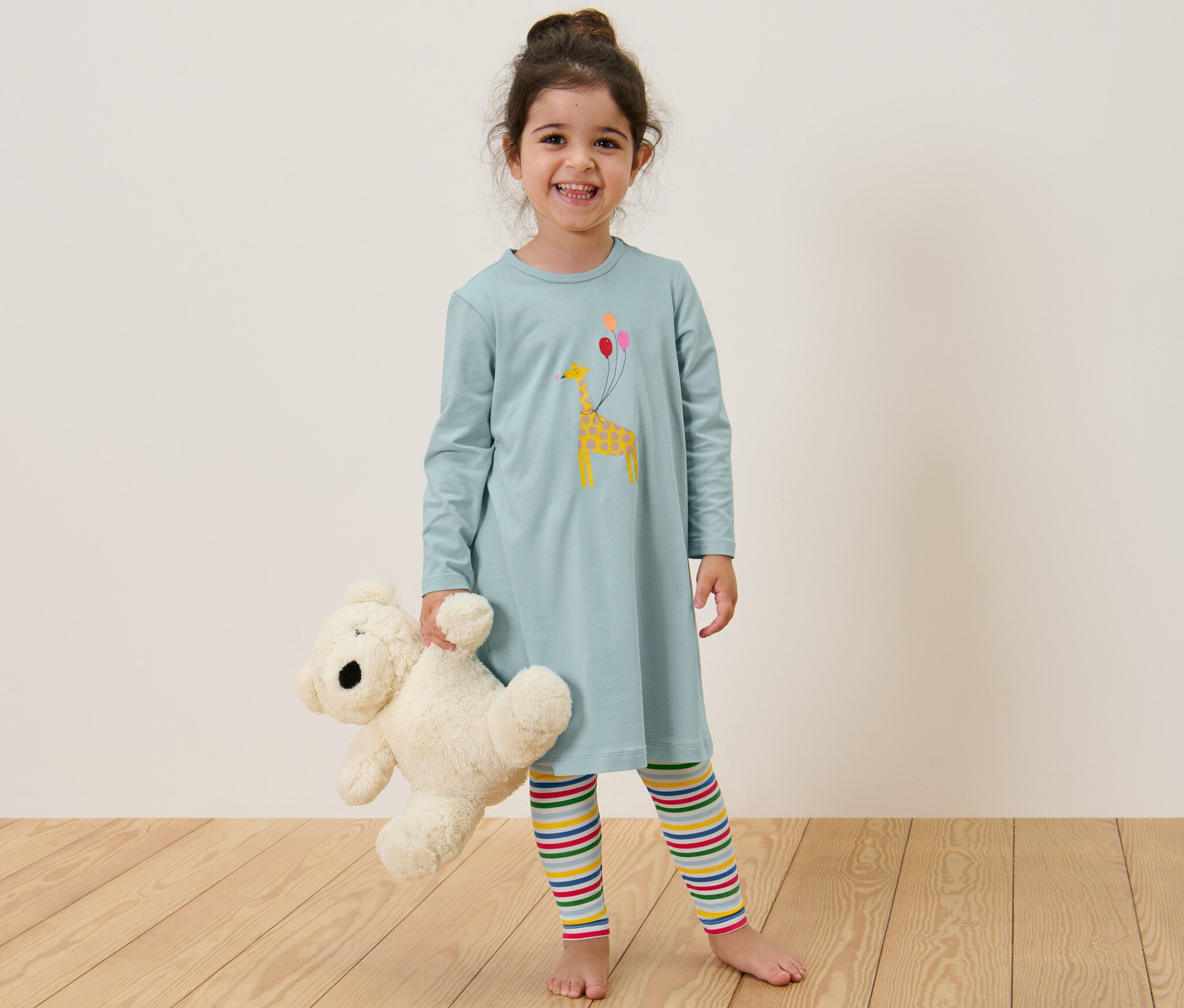 Uśmiechnięta dziewczynka trzyma misia, ubrana w dziecięcą koszulę nocną z legginsami.