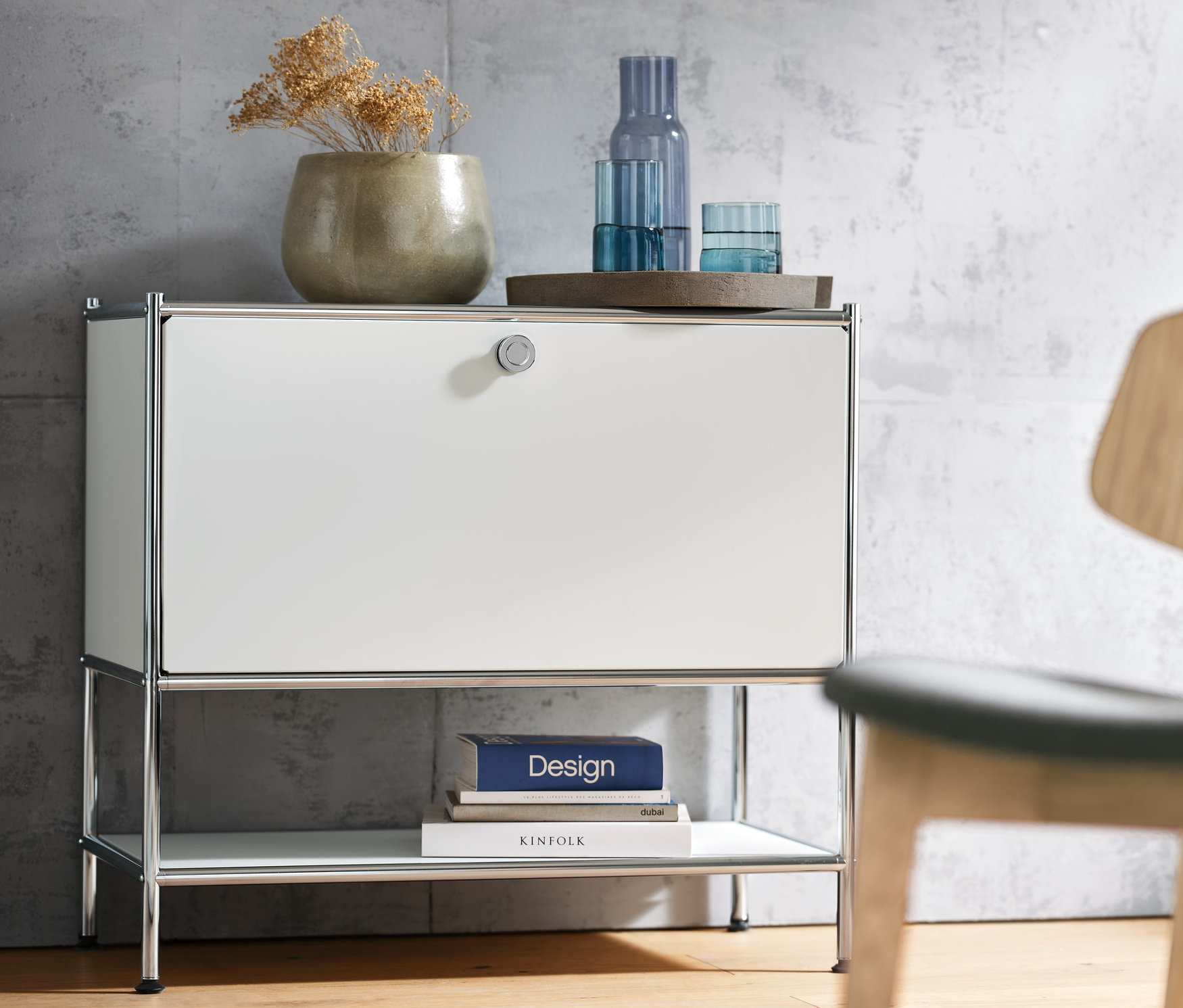 Biała szafka typu sideboard »CN3« z jedną klapą, na której stoi wazon i szklanki. Po prawej krzesło.