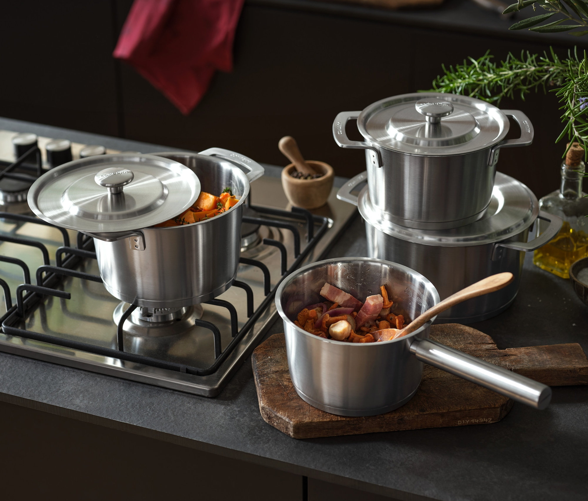 Zestaw garnków ze stali nierdzewnej COMBEKK stoi na kuchence i obok na blacie. W jednym garnku leży drewniana łyżka. W tle widoczne są Ścierki do naczyń, 2 sztuki.