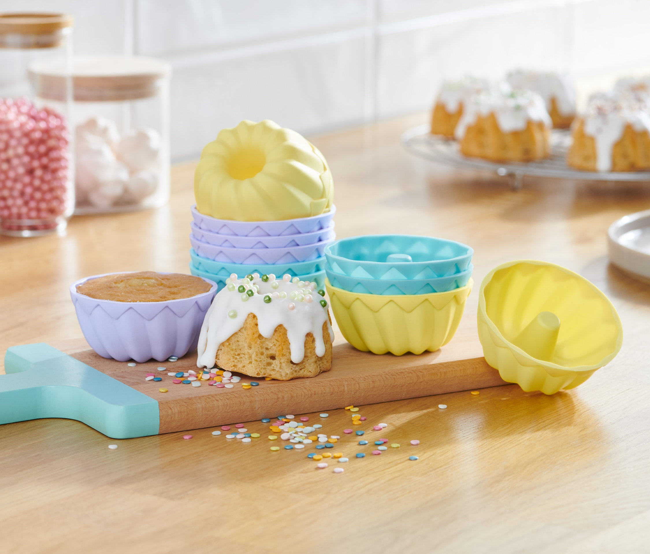 Mini babeczki w kolorowych foremkach na drewnianej desce. W tle słoiki ze słodyczami.