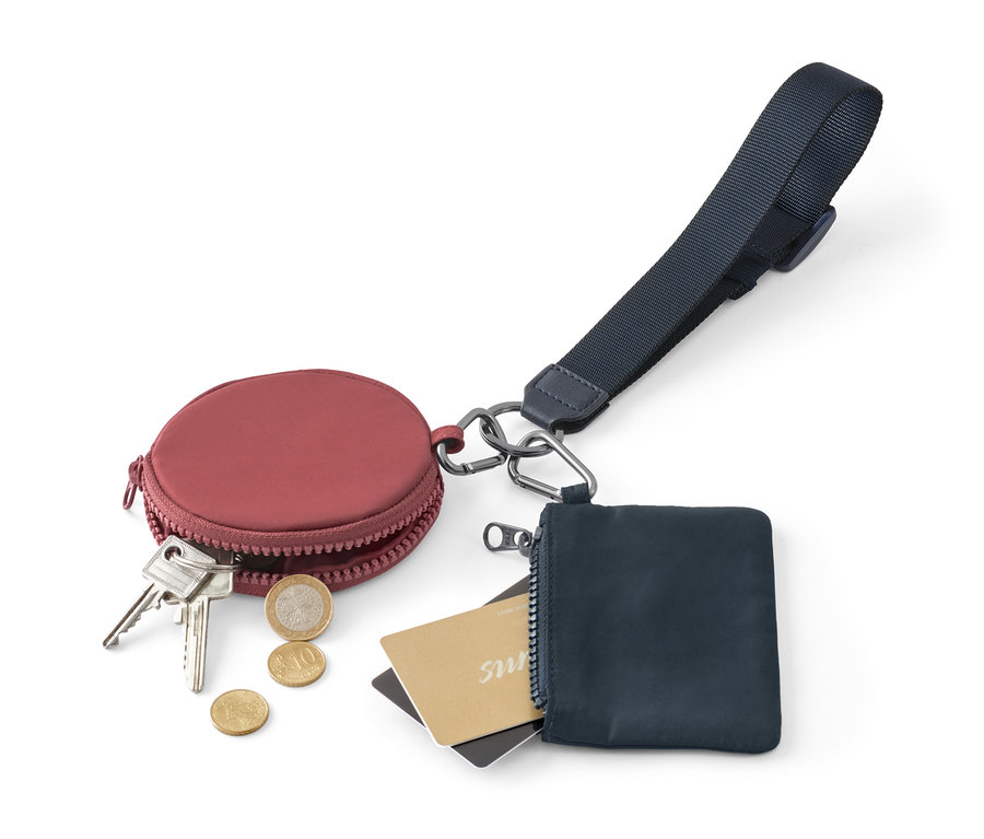 Bordowe, okrągłe etui na monety z kluczami i monetami obok niebieskiego etui na zamek błyskawiczny z kartami i brelokiem-smyczą z kieszonkami do przechowywania.