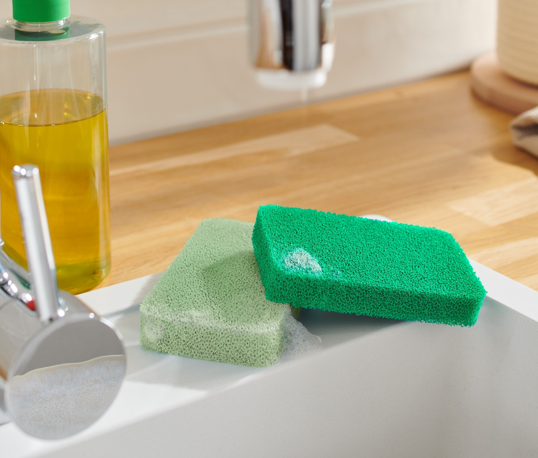 Dwie silikonowe gąbki leżą w białym zlewie obok kranu i butelki ze środkiem czyszczącym.