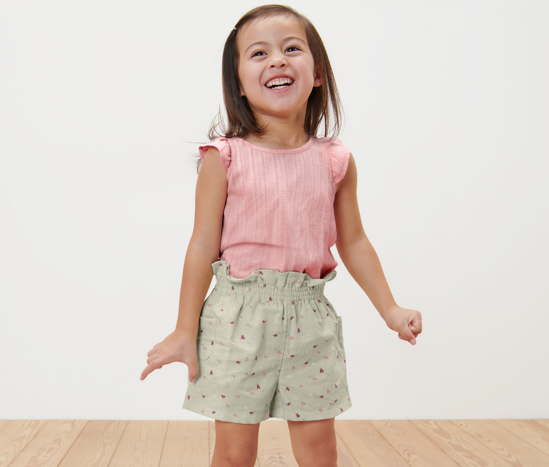 Uśmiechnięta dziewczynka stoi w dziecięcych szortach typu paperbag i różowym topie.