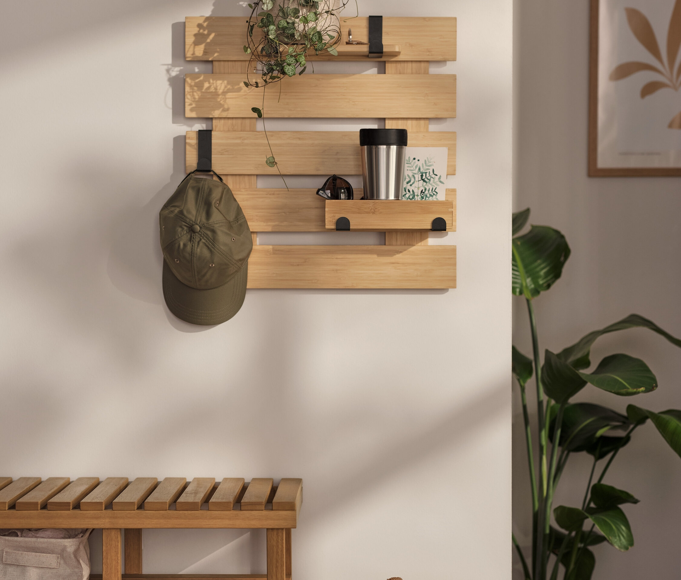 Na ścianie wisi półka z ruchomymi haczykami i podstawą. Na niej stoi roślina, termos i kartka. Na jednym z haczyków wisi czapka z daszkiem.