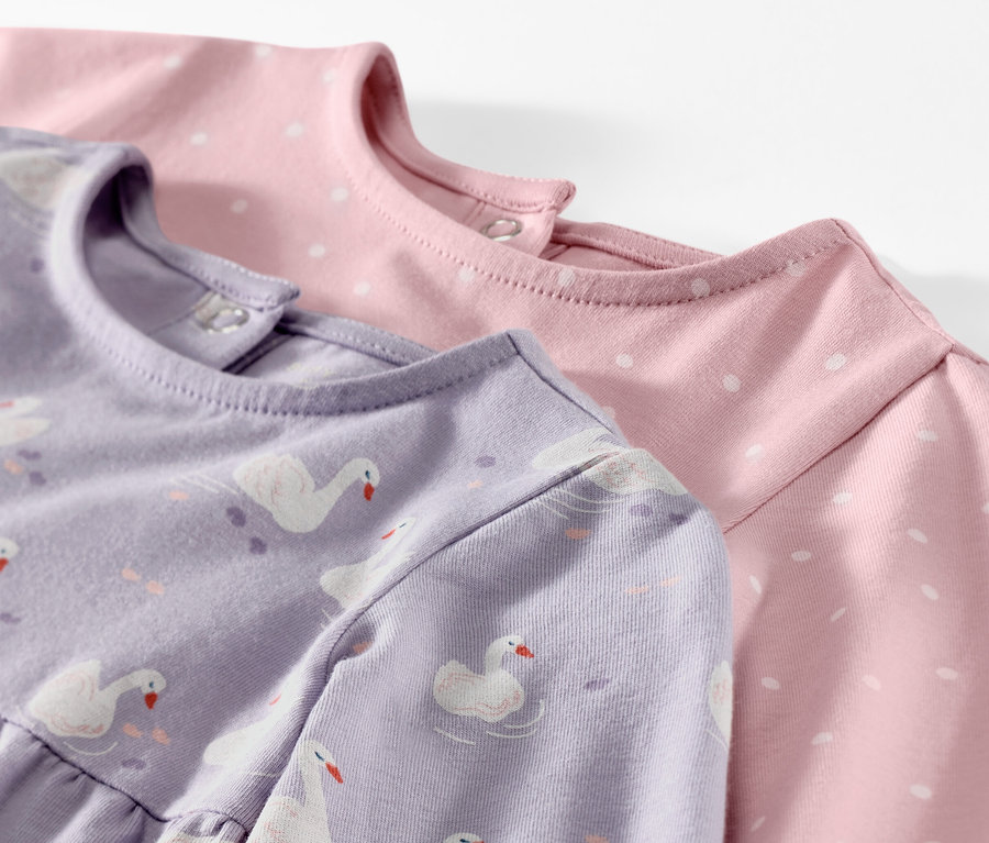 Zbliżenie na dwie dżersejowe sukienki dla niemowląt, jedna liliowa we wzór łabędzi, druga różowa w białe kropki.