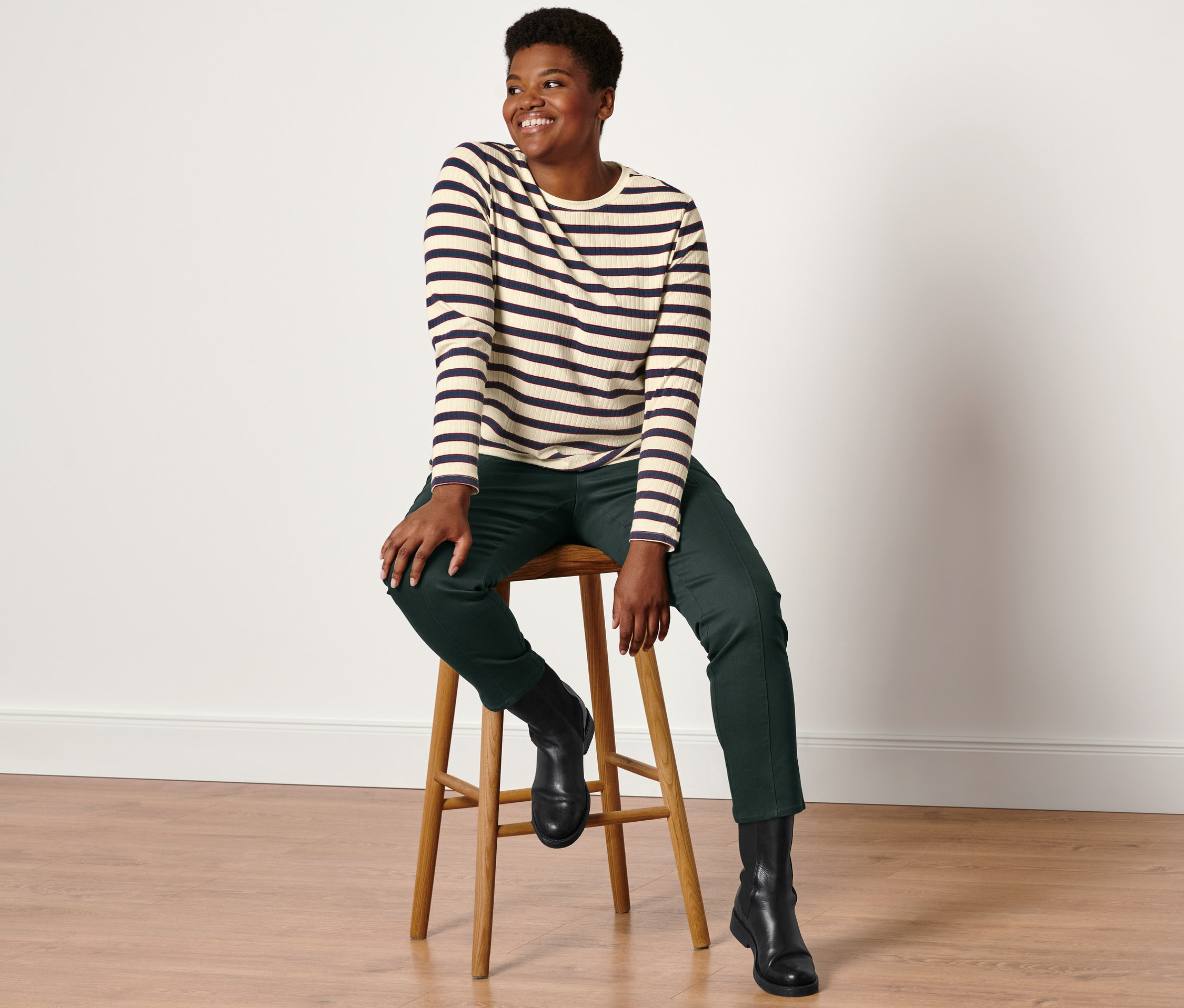 Kobieta siedzi na stołku, ma na sobie trykotową bluzkę z długim rękawem, spodnie z tkaniny twill z wygodnym wykończeniem w pasie w kolorze ciemnozielonym i skórzane buty.