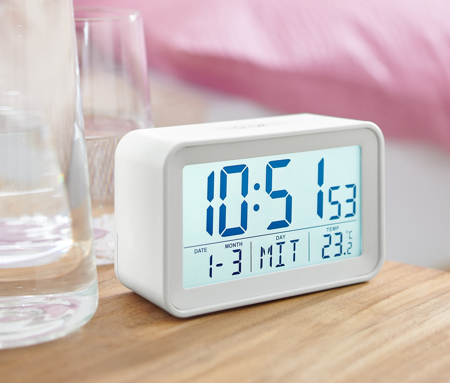 Biały budzik cyfrowy LCD stoi na szafce nocnej obok szklanki wody.