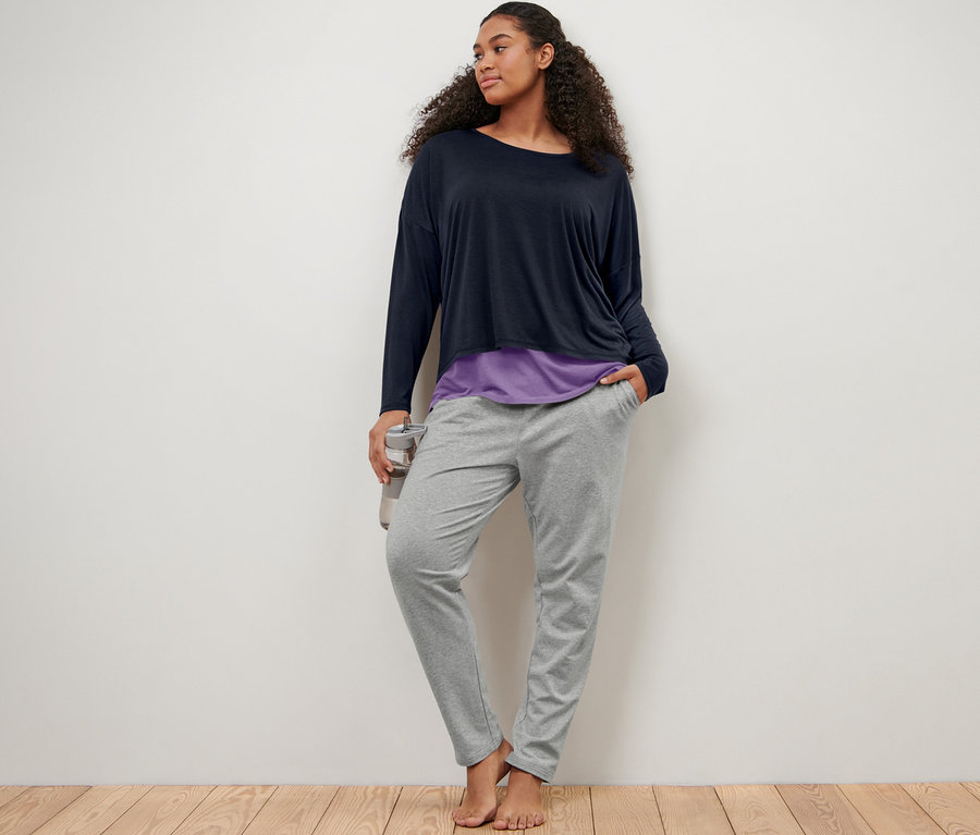 Kobieta opiera się o ścianę, ubrana w szare spodnie dresowe i niebiesko-liliową koszulkę sportową 2 w 1.