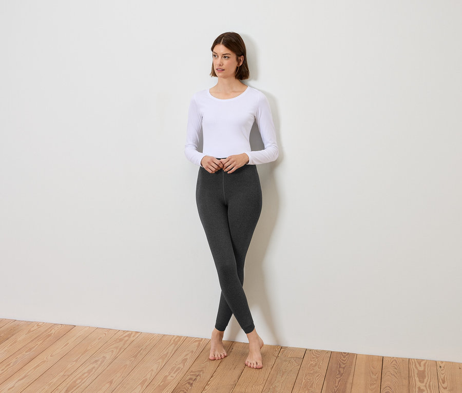 Kobieta stoi ze skrzyżowanymi nogami, ubrana w białą bluzkę z długim rękawem i szare legginsy.