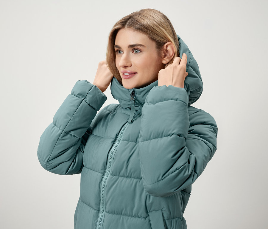 Kobieta poprawia kaptur na turkusowej kurtce puchowej.