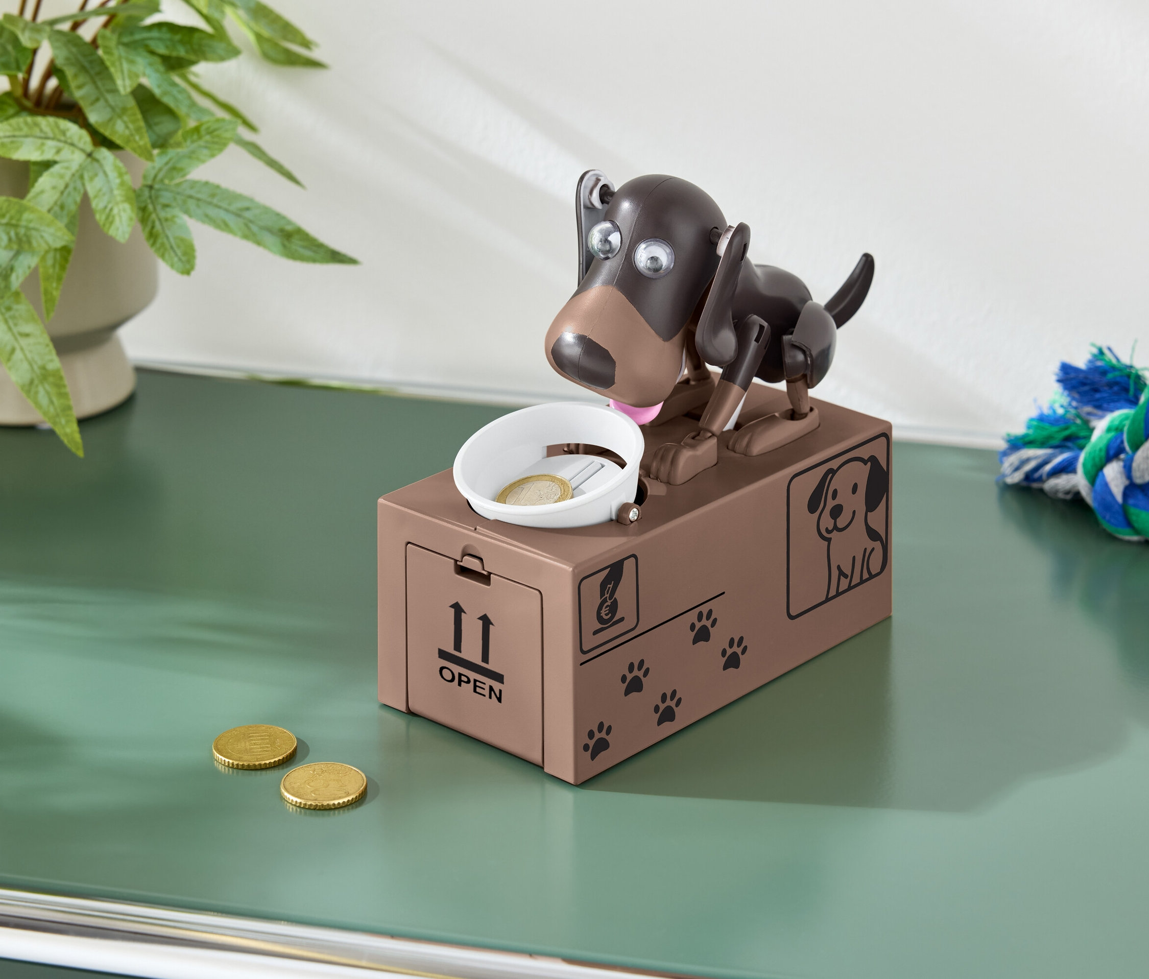 Brązowa skarbonka w kształcie psa z monetą w misce i dwoma monetami na zielonym stole.
