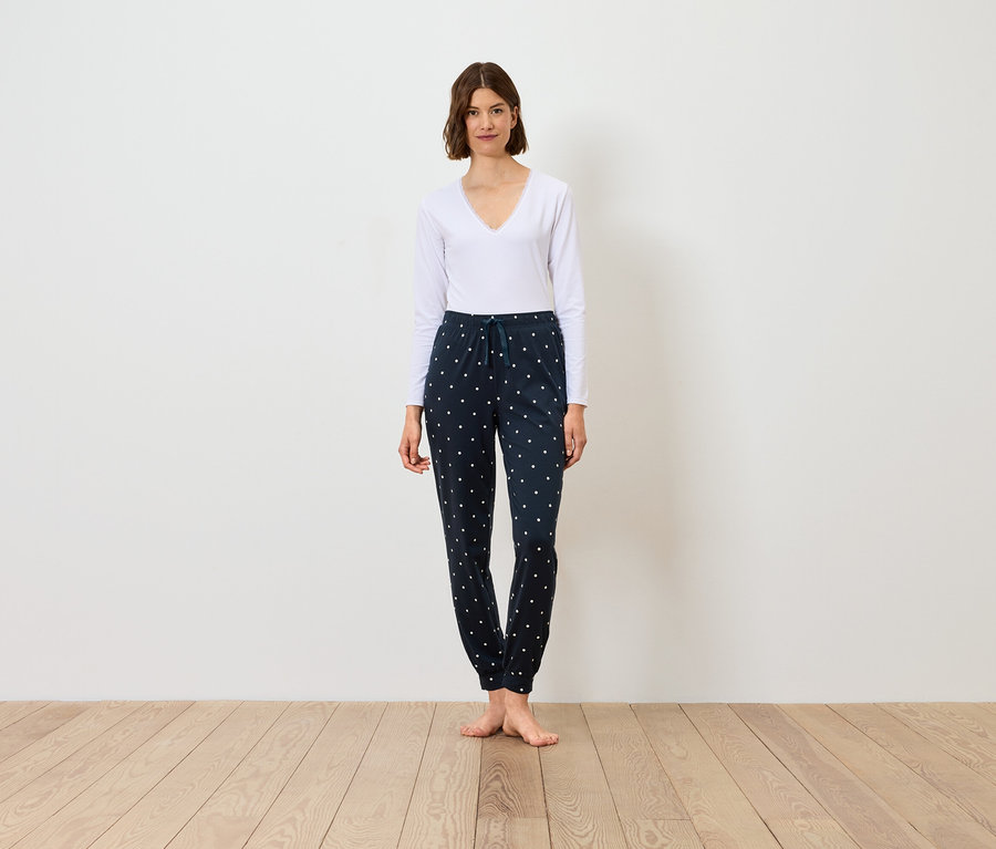 Kobieta stoi, ubrana w białą bluzkę z długim rękawem i granatowe spodnie we wzory.