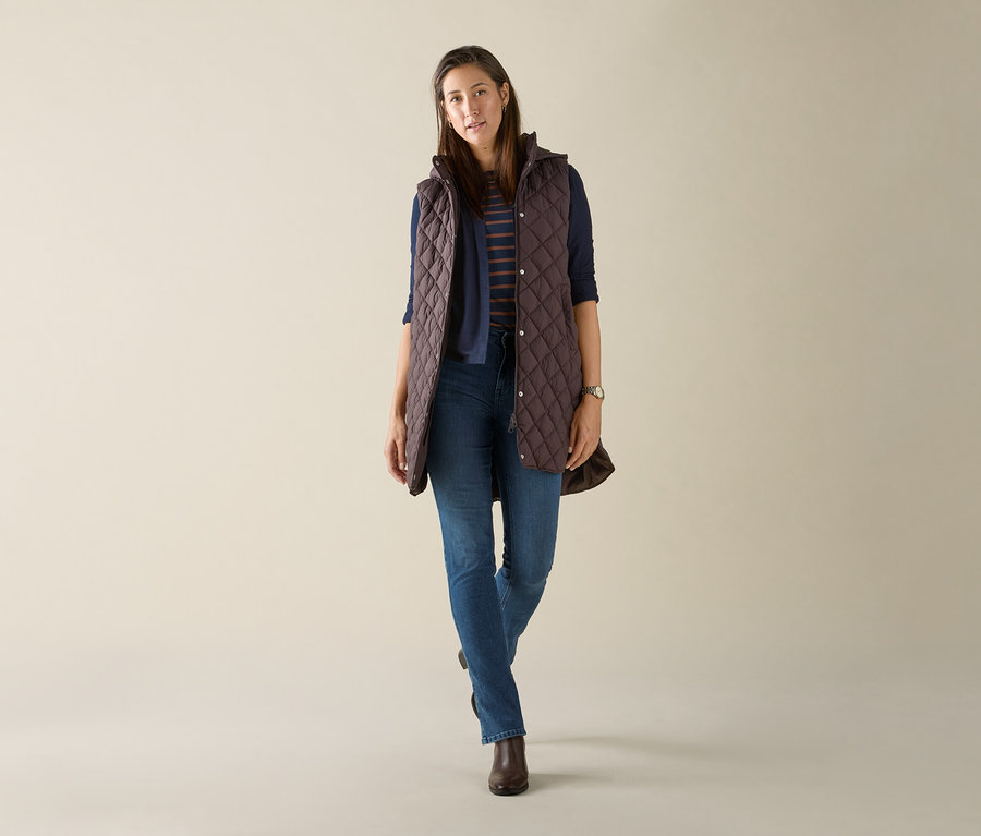 Kobieta ma na sobie długą pikowaną kamizelkę z kapturem w kolorze ciemnobrązowym.