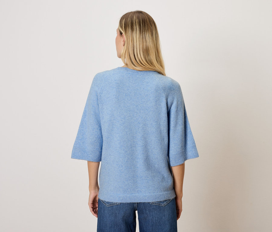 Kobieta o blond włosach stoi tyłem do kamery, ma na sobie niebieski top i dżinsy.