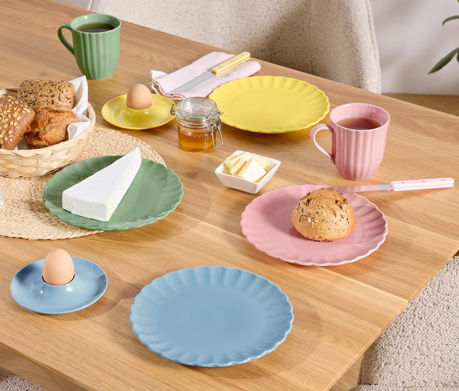 Na drewnianym stole znajdują się kolorowe talerze, filiżanki i miseczki z jedzeniem, w tym chleb w koszyku, jajko w kieliszku i kawałek sera.