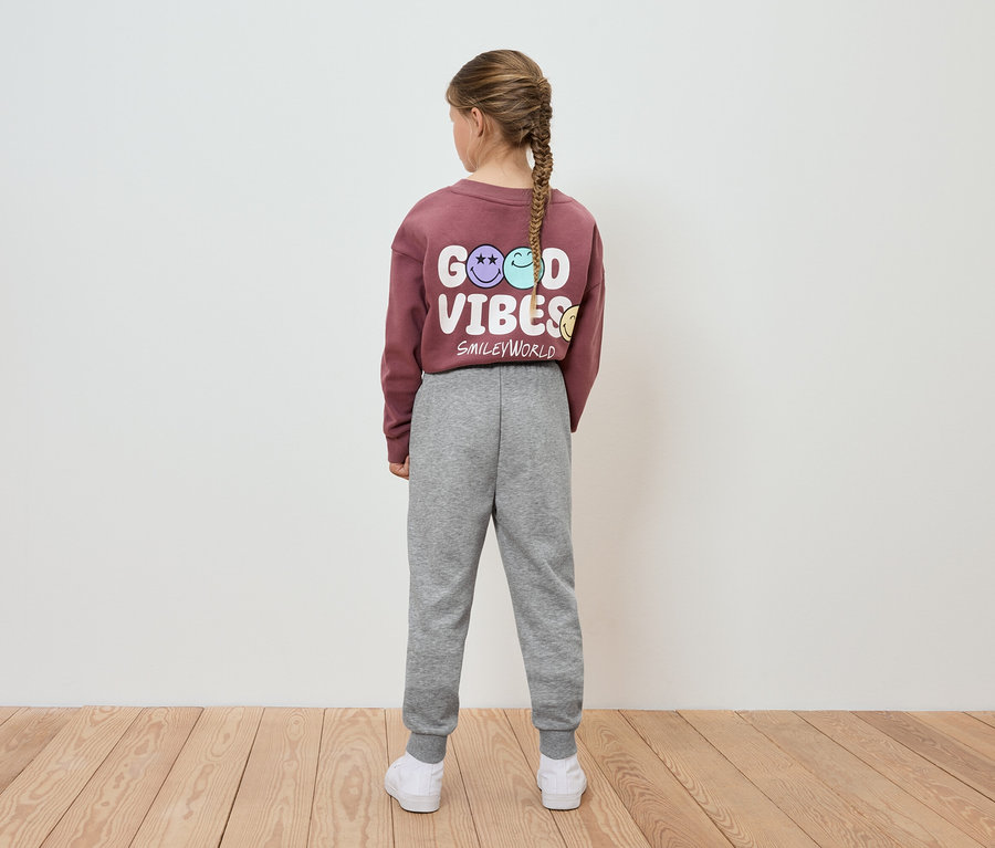 Dziewczynka w bordowej bluzie z napisem Good Vibes i szarych spodniach dresowych stoi tyłem do aparatu.