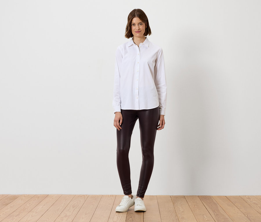 Kobieta w białej koszuli i brązowych legginsach stoi na drewnianej podłodze.