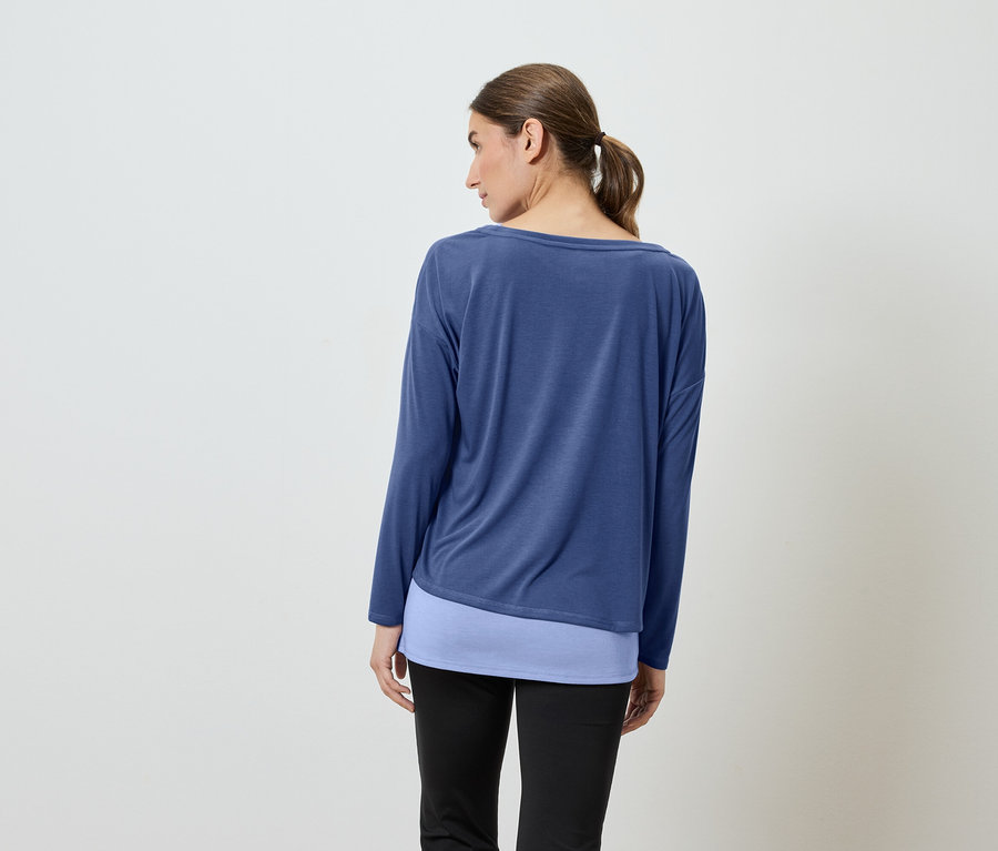 Kobieta tyłem do kamery ma na sobie niebieską koszulę z długim rękawem i czarne spodnie.