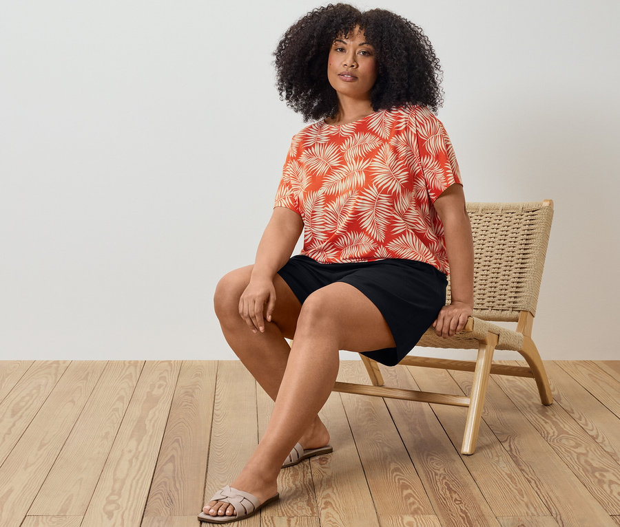 Kobieta siedzi na krześle, ma na sobie wzorzystą bluzkę i czarne szorty z dżerseju.