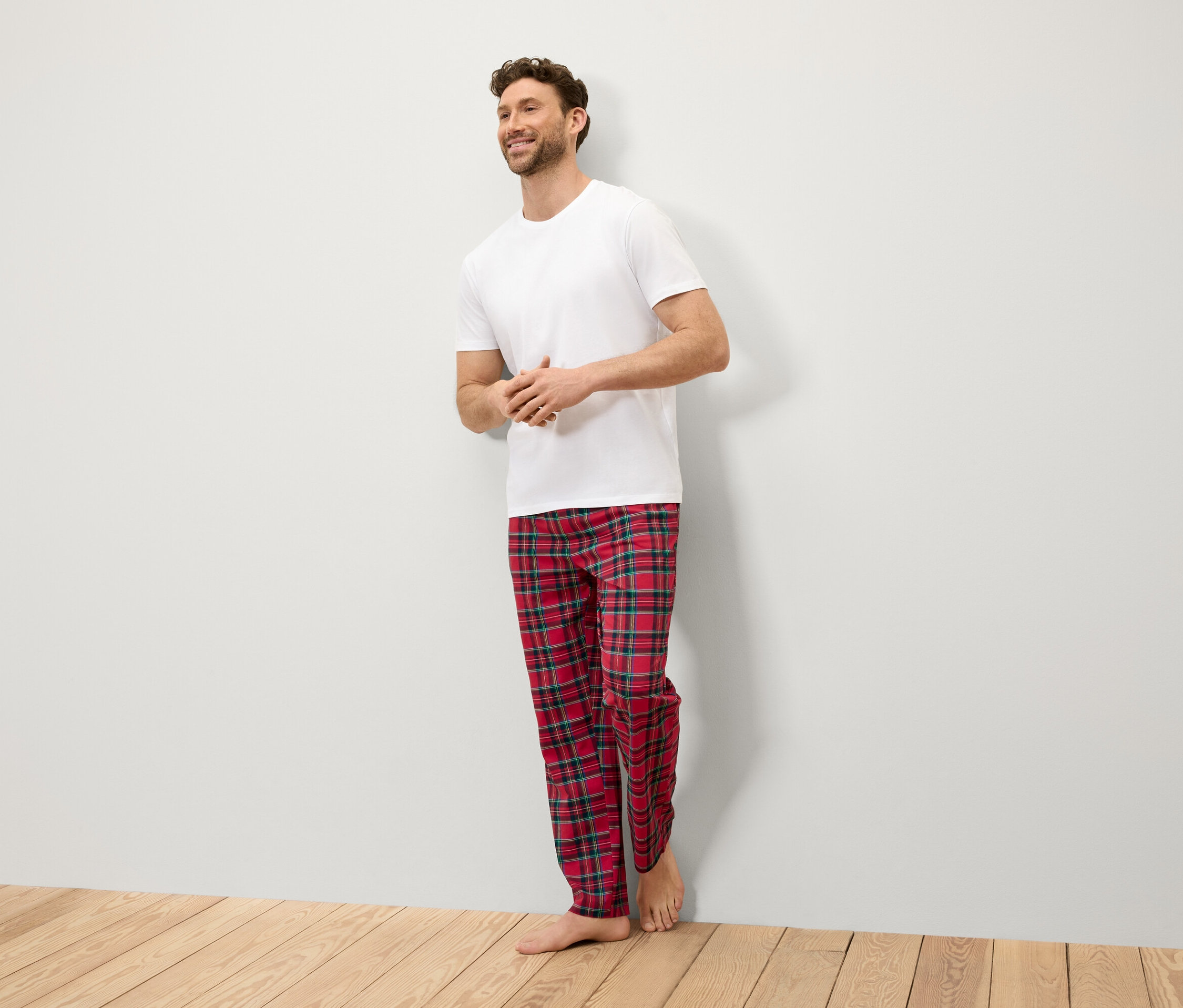 Uśmiechnięty mężczyzna stoi przy ścianie w białej koszulce i czerwonych spodniach w kratę.
