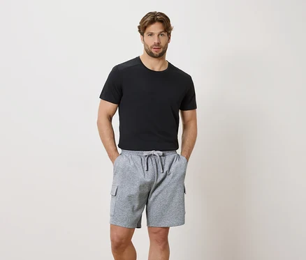 Mężczyzna z rękami w kieszeniach szarych szortów ze sznurkiem i czarnym T-shirtem z krótkim rękawem.