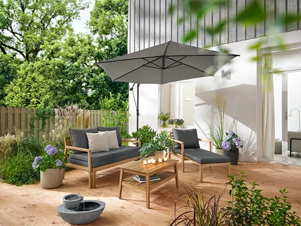 Na drewnianym tarasie stoją rozsuwany fotel ogrodowy, rozsuwana sofa ogrodowa, ogrodowy stolik kawowy (ok. 80 x 50 cm) i solarna fontanna ogrodowa. Parasol Leco w kolorze antracytowym zapewnia cień. 2 poduszki ogrodowe o strukturze lnu i 6 akumulatorowych lampek LED do użytku na zewnątrz zdobią otoczenie.