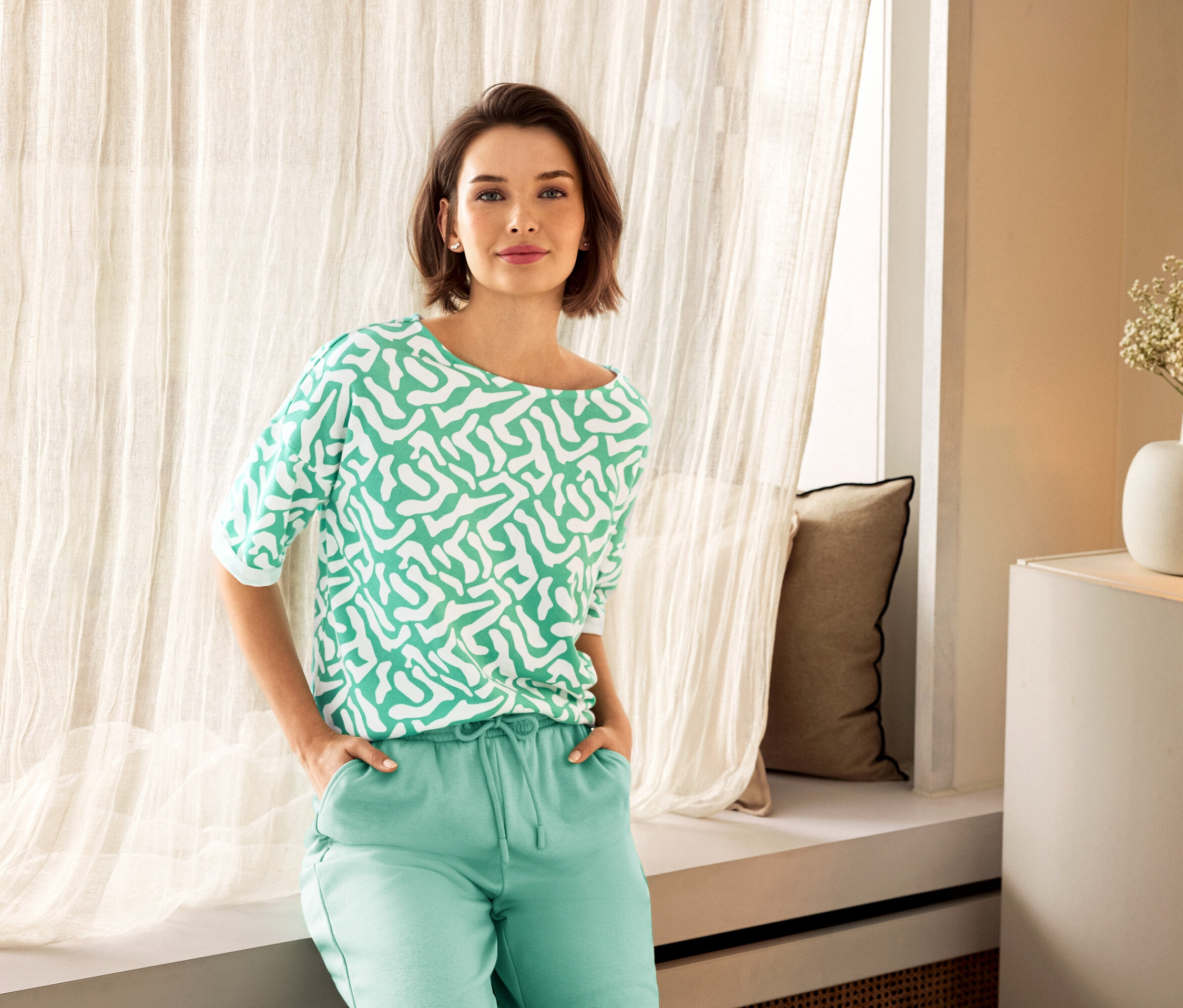 Kobieta w zielonych spodniach i wzorzystej bluzce pozuje obok okna. W tle widać wazon.