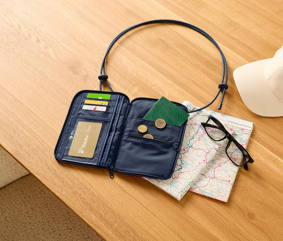 Otwarty, granatowy portfel podróżny z kartami, paszportem i monetami na drewnianym stole, obok mapy, czarnych okularów i białej czapki.