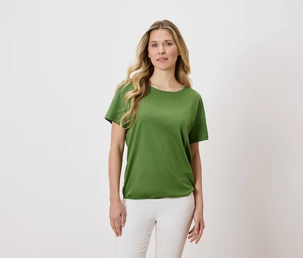 Kobieta o blond, falistych włosach ma na sobie zielony T-shirt i białe spodnie na jasnym tle.