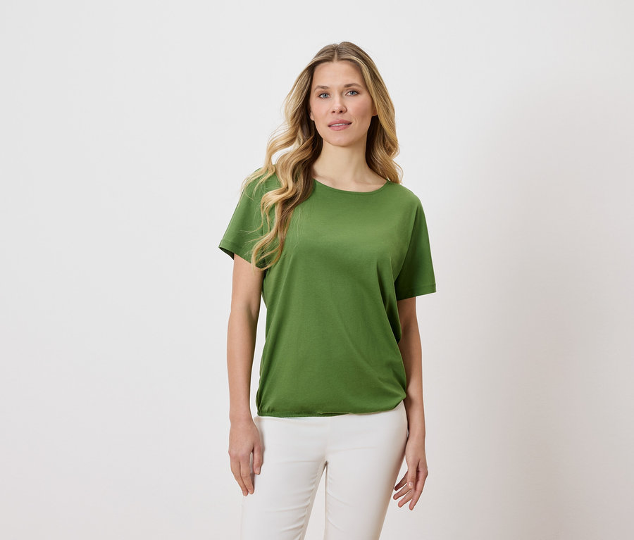 Kobieta o blond, falistych włosach ma na sobie zielony T-shirt i białe spodnie na jasnym tle.