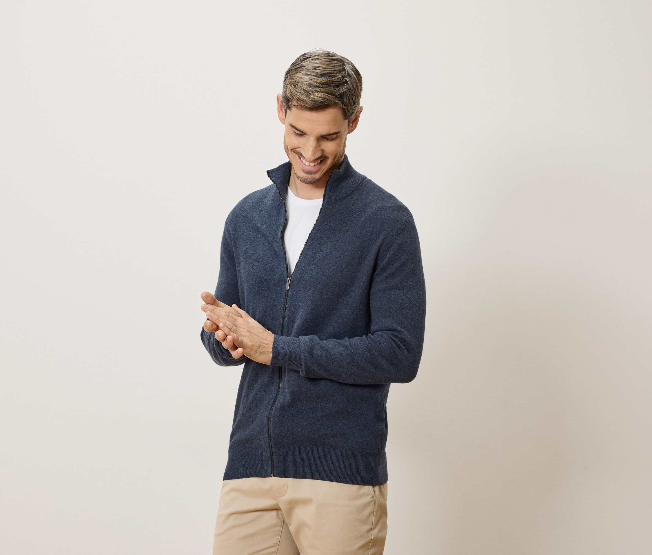 Uśmiechnięty mężczyzna pociera ręce. Ma na sobie granatowy kardigan i beżowe spodnie.