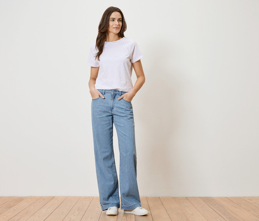 Kobieta w białej koszulce i niebieskich dżinsach stoi na tle białej ściany.