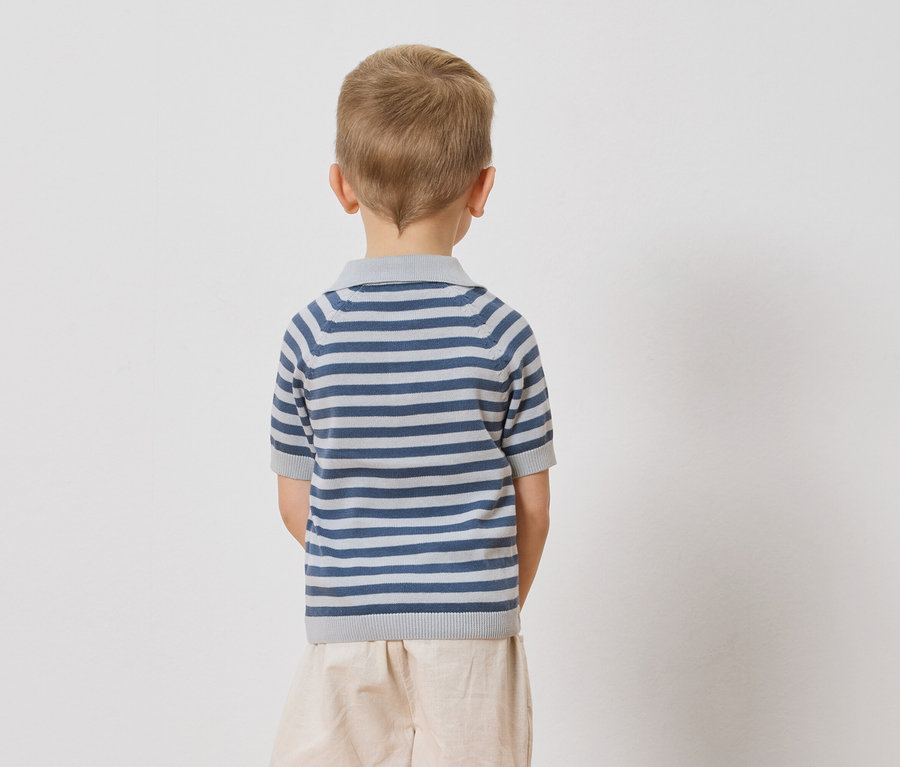 Widok z tyłu dziecka z blond włosami ubranego w polo w niebiesko-białe paski i beżowe szorty na białym tle.