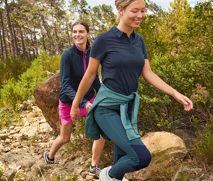 Dwie kobiety wędrują po kamienistej ścieżce, kobieta z przodu ma na sobie granatową koszulkę polo, zielone legginsy i sweter przewiązany w pasie.