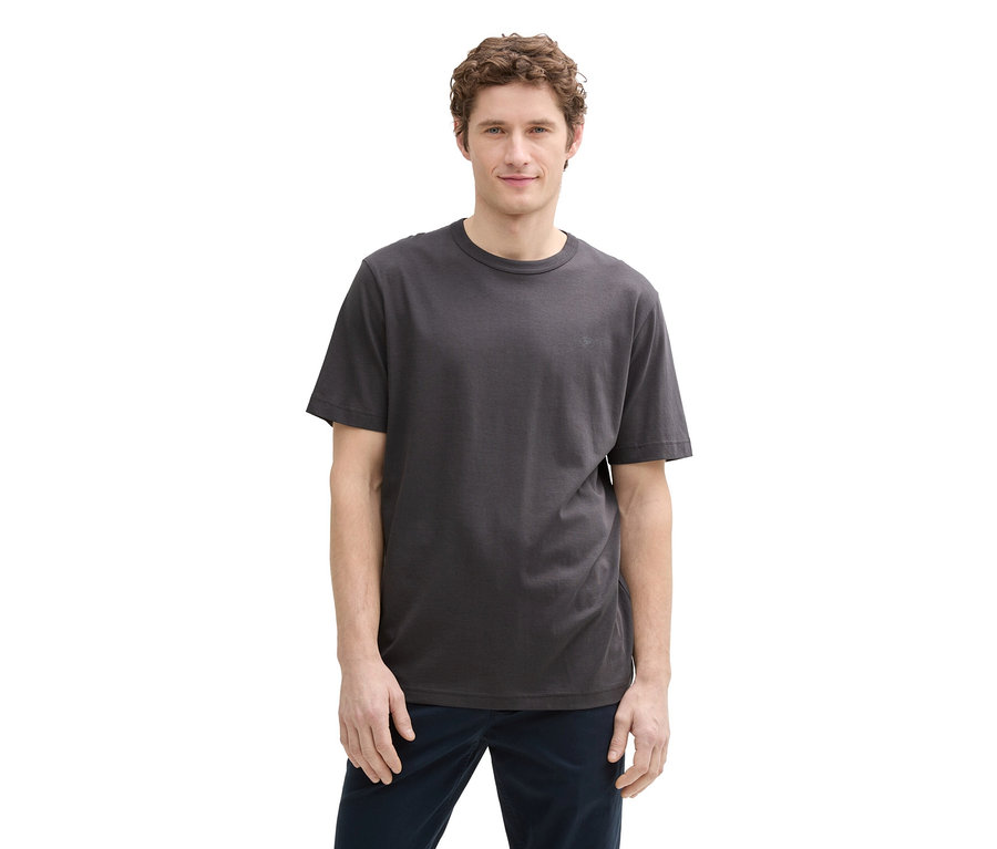 Mężczyzna o kręconych włosach ma na sobie szary T-shirt i niebieskie spodnie, stoi na białym tle.