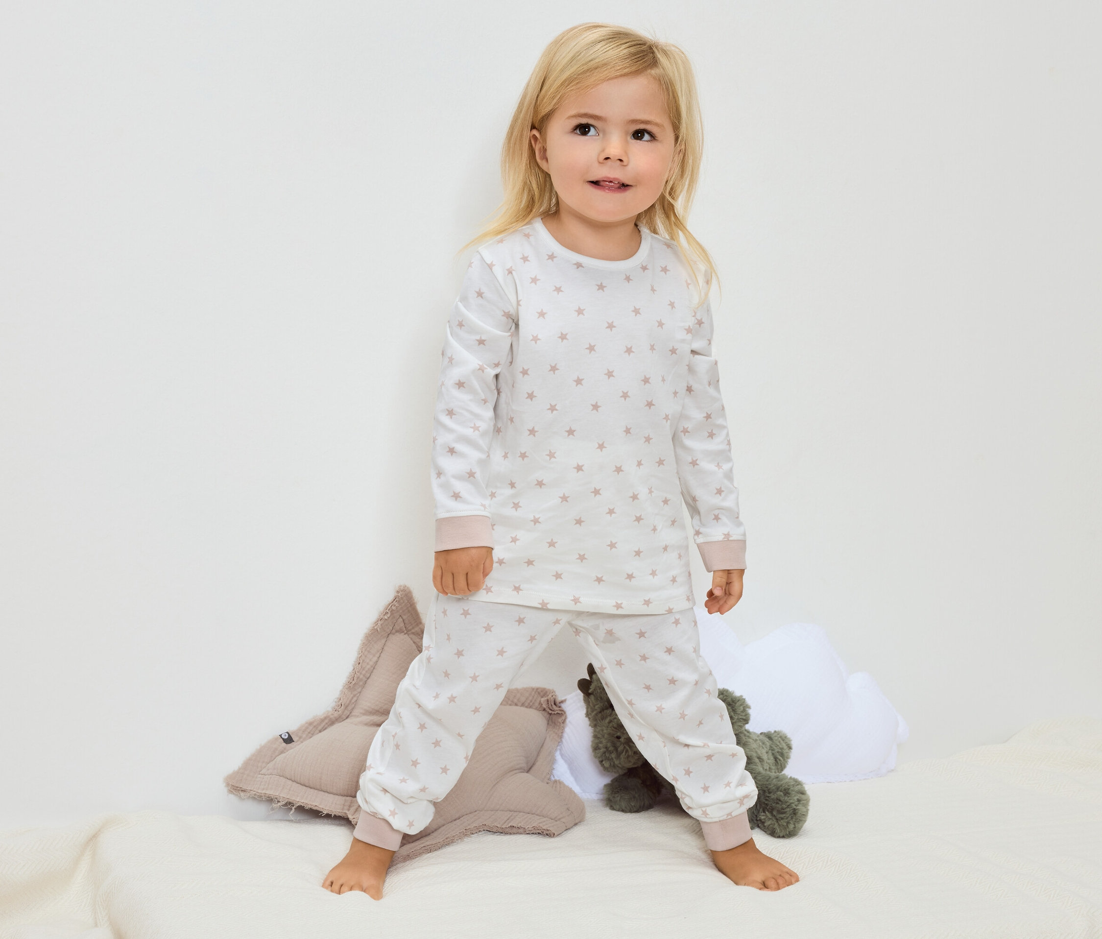 Mała blondynka stoi boso na kocu, ubrana w białą piżamę w gwiazdki.