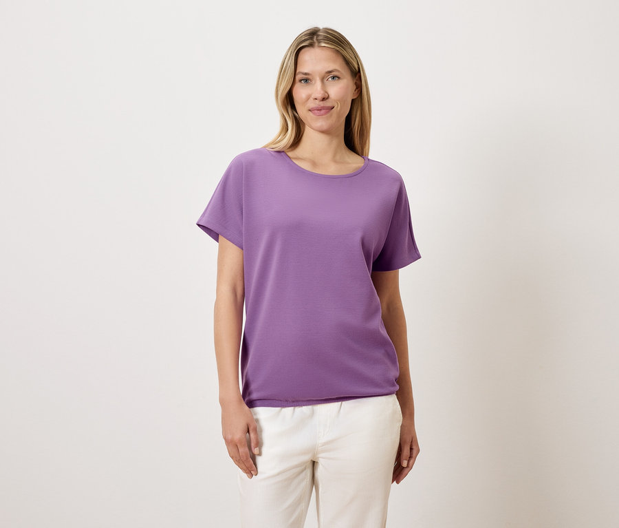 Kobieta o blond włosach ma na sobie fioletowy T-shirt i białe spodnie.