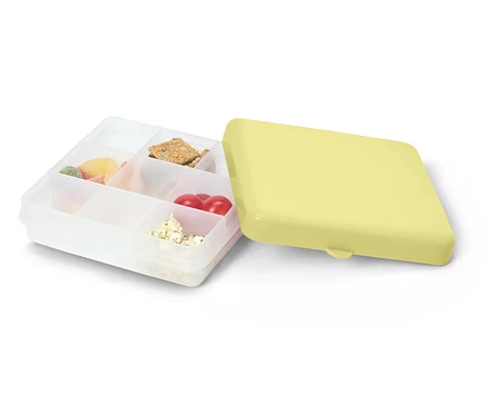 Otwarty, przezroczysty pojemnik na lunch z żółtą pokrywką. Wypełniony słodyczami, krakersami, pomidorami i serem.