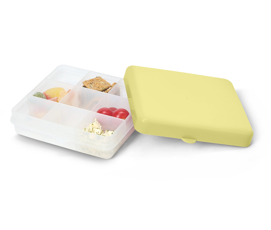Otwarty, przezroczysty pojemnik na lunch z żółtą pokrywką. Wypełniony słodyczami, krakersami, pomidorami i serem.