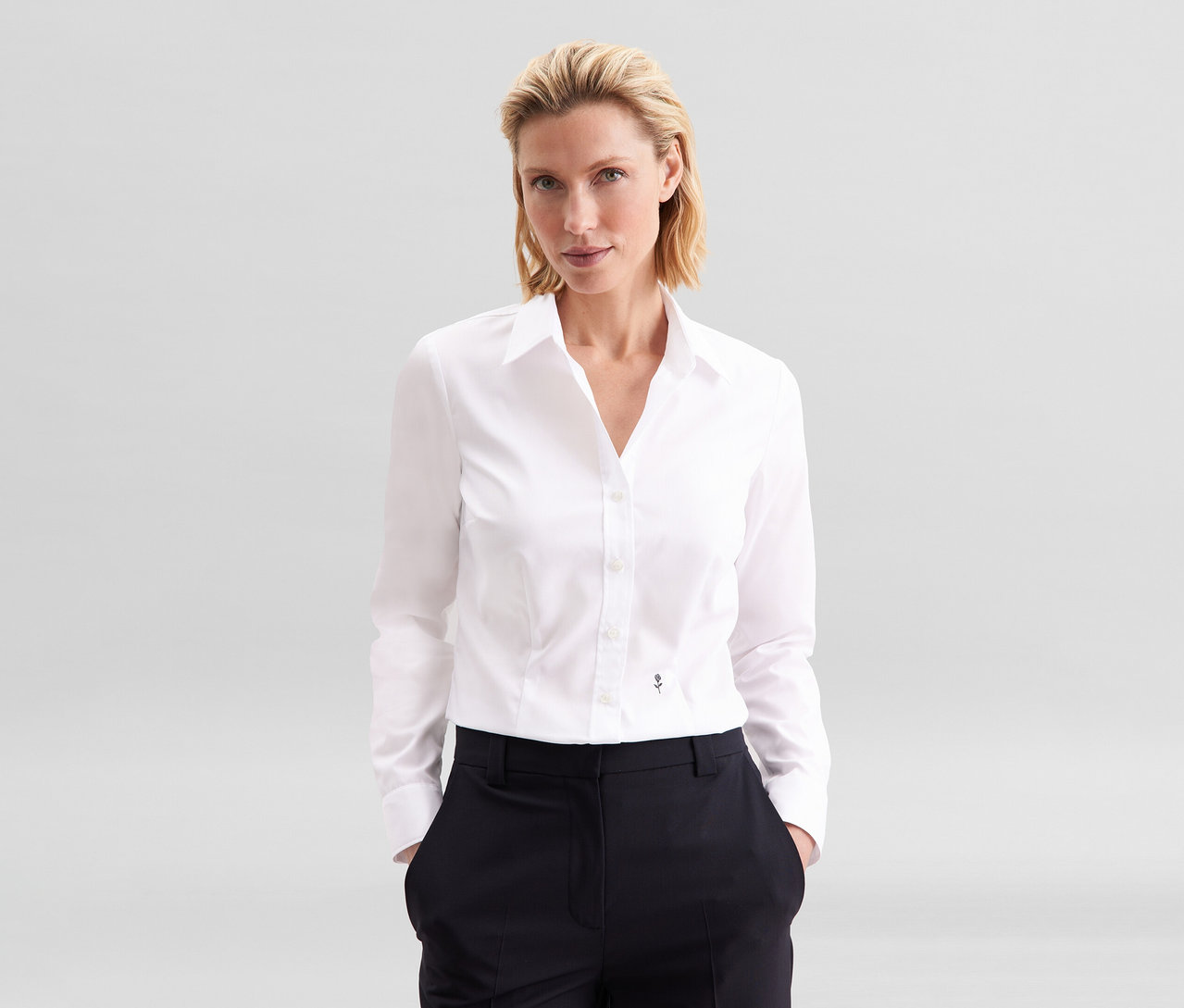 Blondynka w białej bluzce Seidensticker Slim Fit non iron i ciemnych spodniach, z rękami w kieszeniach.