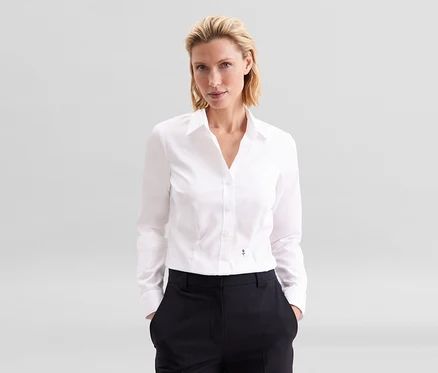 Blondynka w białej bluzce Seidensticker Slim Fit non iron i ciemnych spodniach, z rękami w kieszeniach.