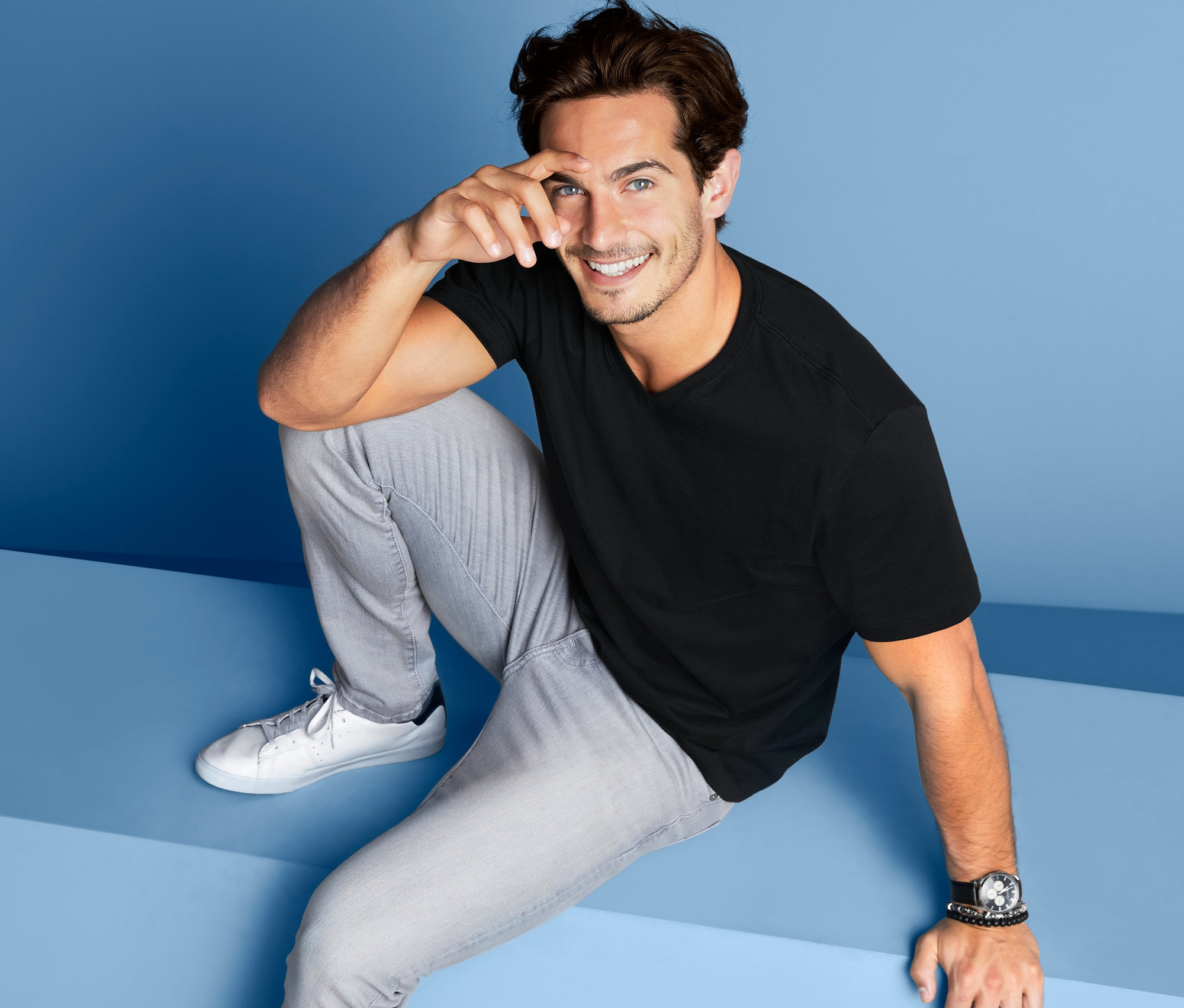 Mężczyzna siedzi na niebieskim tle i ma na sobie czarny T-shirt z dekoltem w serek z zestawu 2 sztuk.