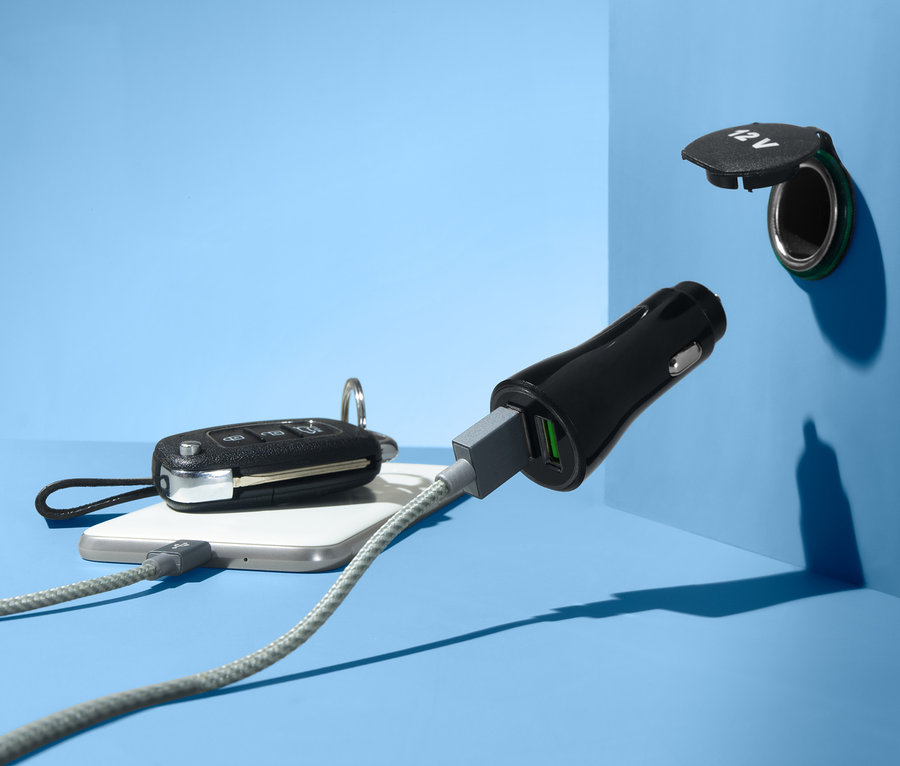 Na niebieskim tle widoczna jest ładowarka samochodowa z portem USB podłączona do gniazda 12V. Kabel jest podłączony do telefonu komórkowego.