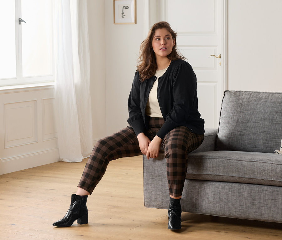 Kobieta siedzi na szarej sofie, ubrana w kremową koszulkę, spodnie stretchowe w kratę i sweter z delikatnej dzianiny z bufiastymi rękawami.